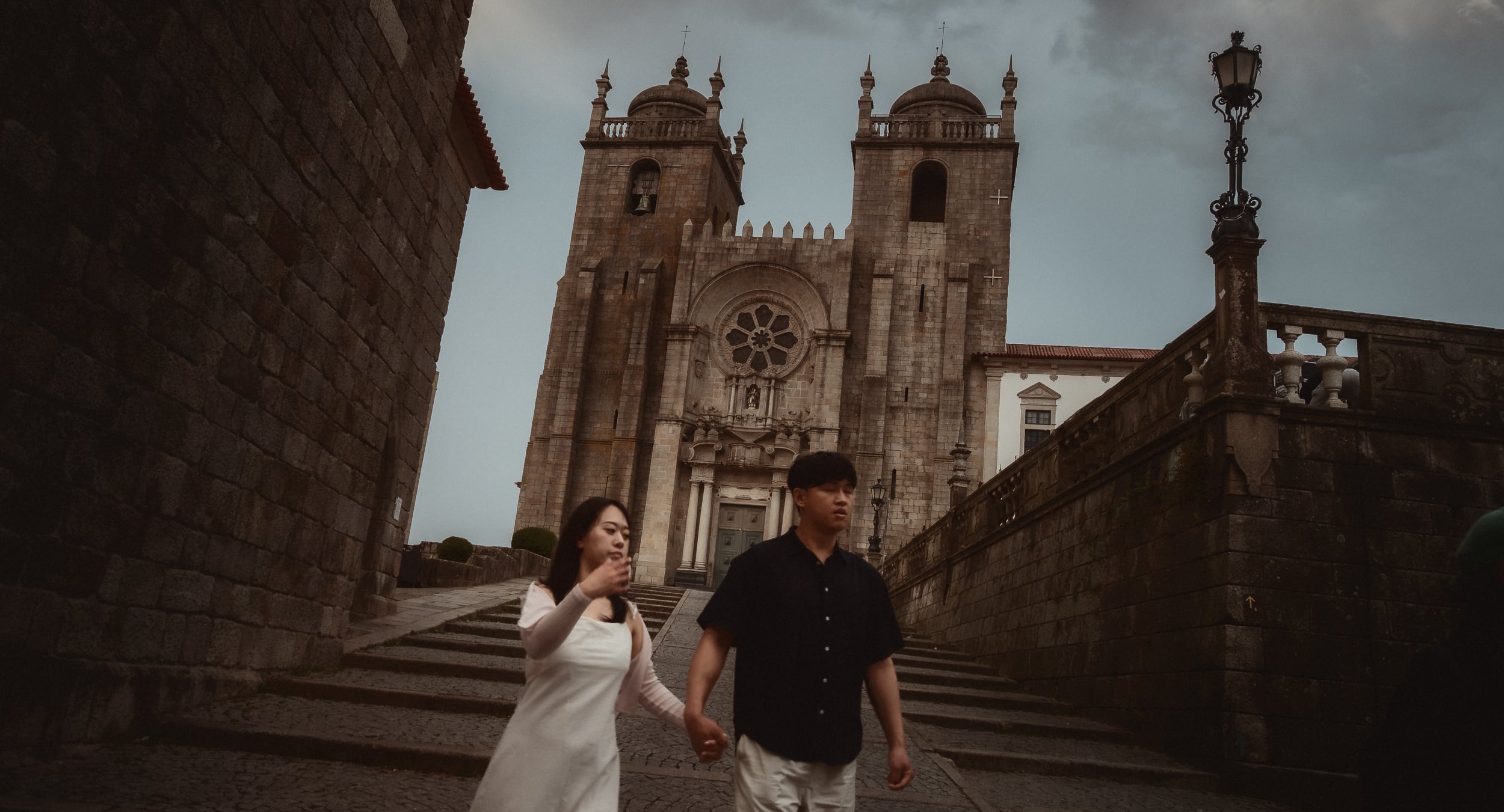 Una pareja cogida de la mano baja las escaleras delante de la catedral de Oporto, con el cielo gris.